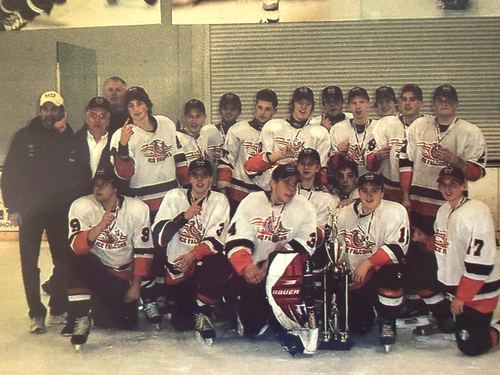 Team in white jerseys kneeling and standing around a trophy on the ice